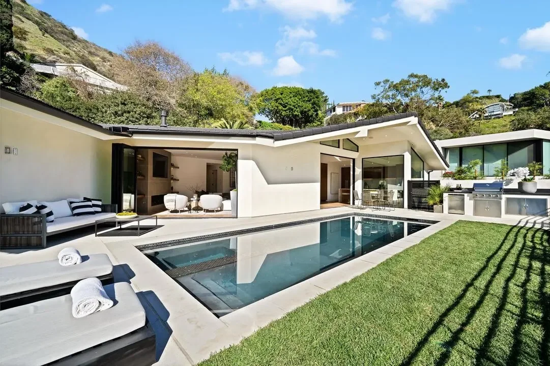 Modern house with outdoor swimming pool, lounge chairs, outdoor kitchen, and patio area, surrounded by hills and greenery under a partly cloudy sky.
