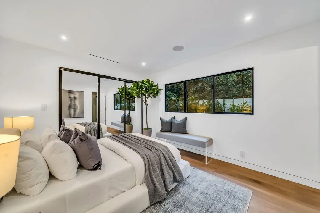 Modern bedroom with white walls, large windows, a white bed with gray and purple pillows, a gray throw, wooden floors, a bench with two gray pillows, and indoor plants.