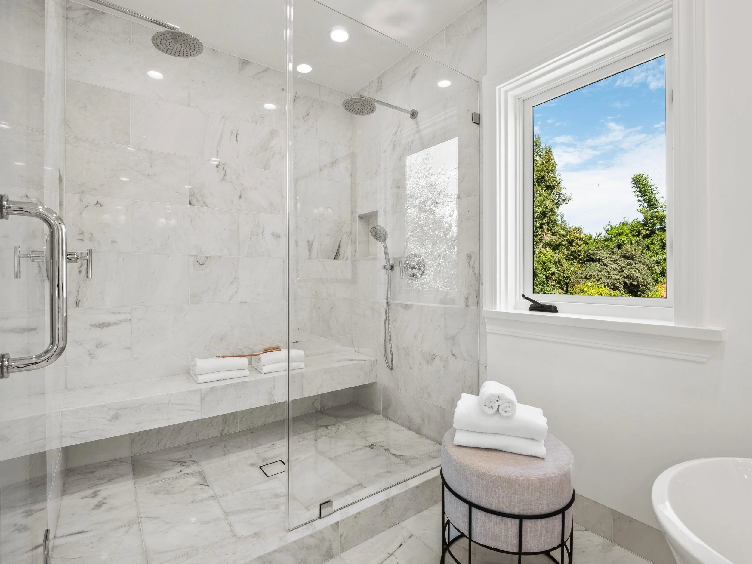 Hancock Park primary shower with floor-to-ceiling marble tile, frameless glass enclosure, built-in bench, dual showerheads, and large window with treetop views.