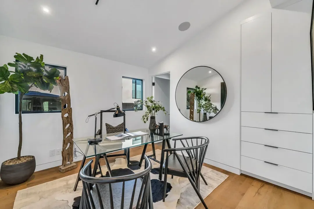 Modern dining room with white walls, wooden flooring, a round mirror, black and glass table, four black chairs, tall white cabinets, green indoor plants, and windows letting in natural light.