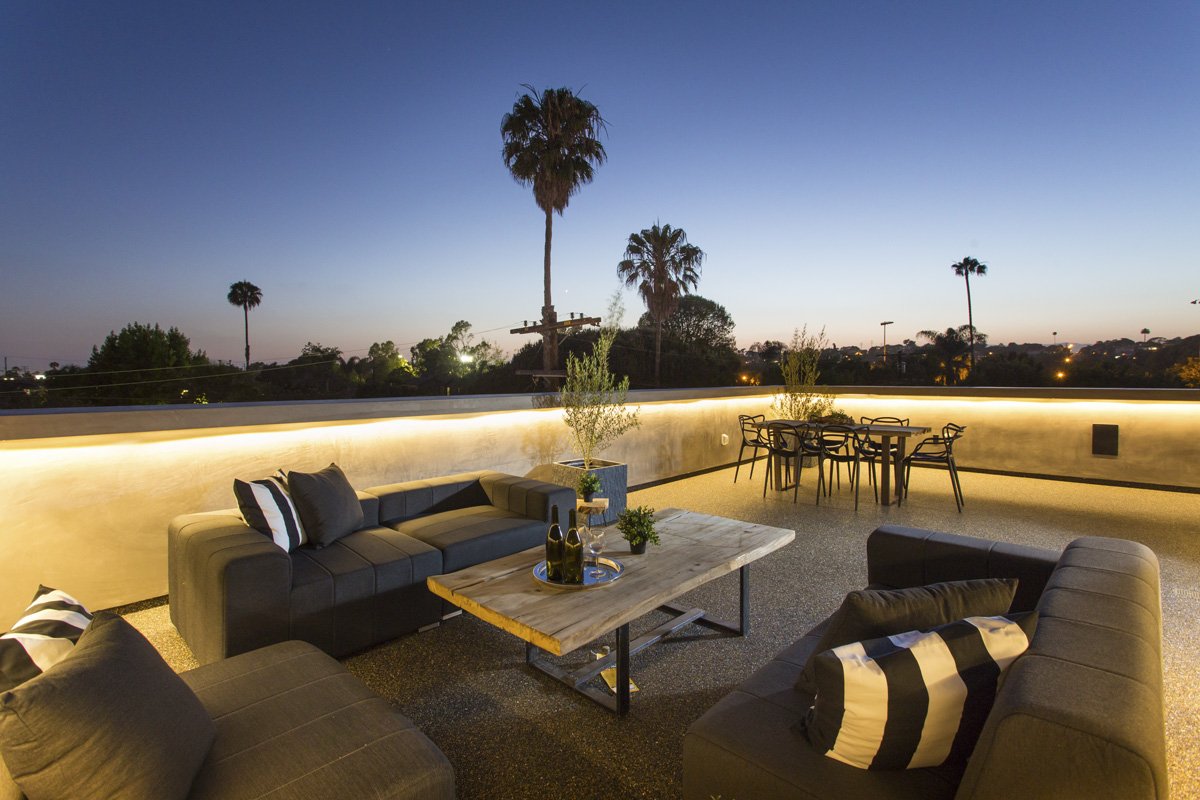 Rooftop dining terrace at sunset with built-in perimeter lighting, contemporary outdoor table and chairs, and tree canopy skyline views.