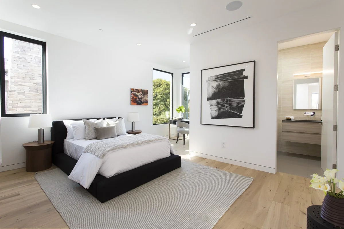 Contemporary guest bedroom with black upholstered bed frame, layered neutral textiles, large corner windows, and ensuite bath access.