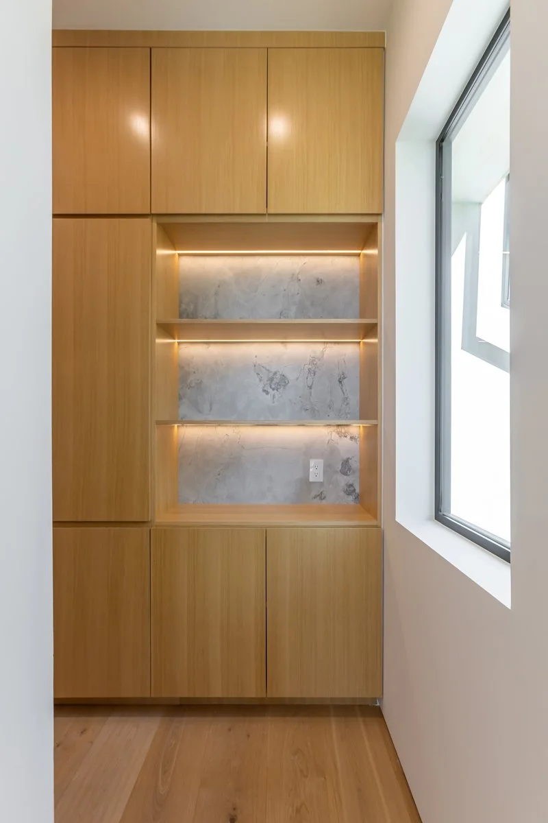 Custom Mar Vista built-in cabinetry with light wood panels, integrated open shelving with warm LED accent lighting, stone back panels, and minimalist modern detailing by Heart Construction.