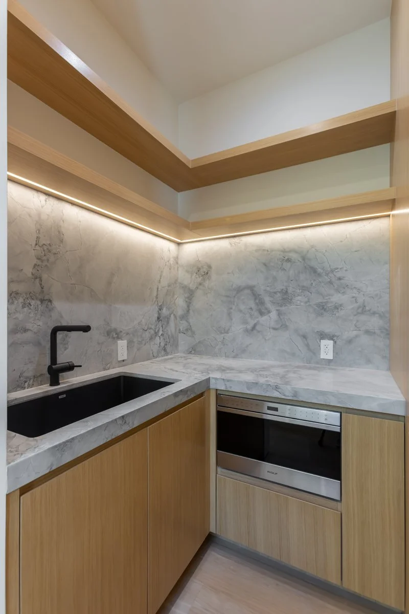 Modern Mar Vista kitchenette with marble backsplash and countertops, custom light wood cabinetry, floating wood shelves with integrated LED lighting, black undermount sink, and built-in microwave in a contemporary custom home.