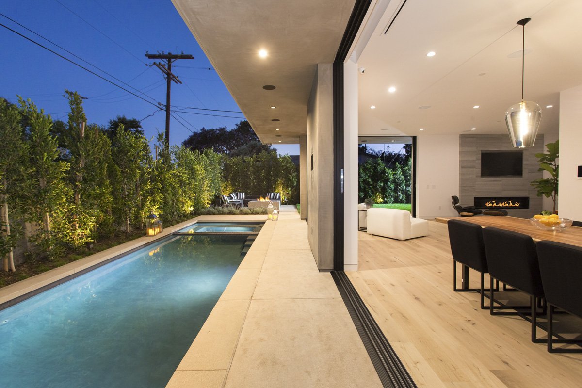 Open-plan dining and living space with flush sliding glass system opening directly to pool deck, continuous flooring transition, and perimeter hedge privacy.