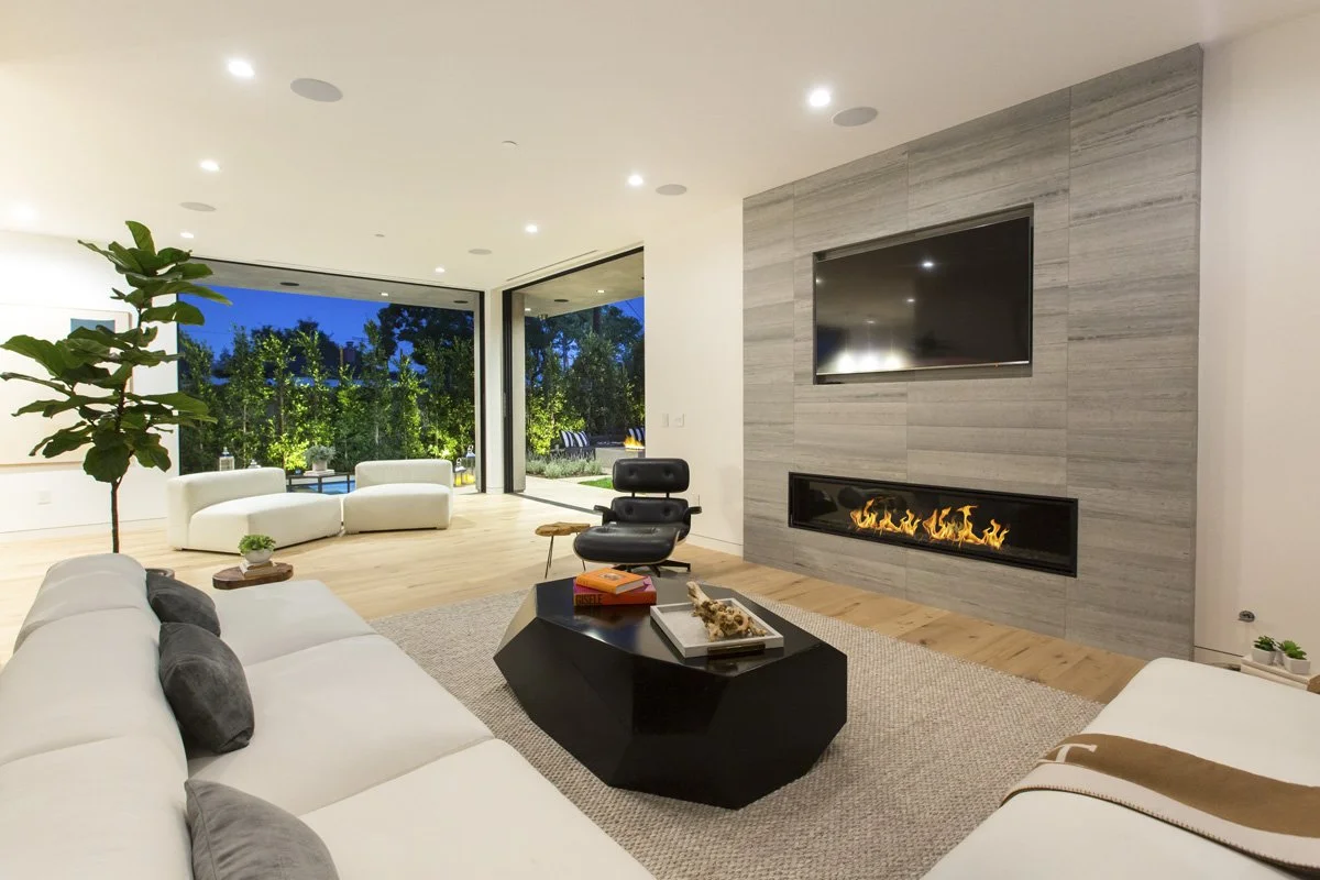 Open-concept living room with wide-plank oak flooring, full-height sliding glass walls, large-format stone fireplace surround, and seamless indoor-outdoor connection at dusk.