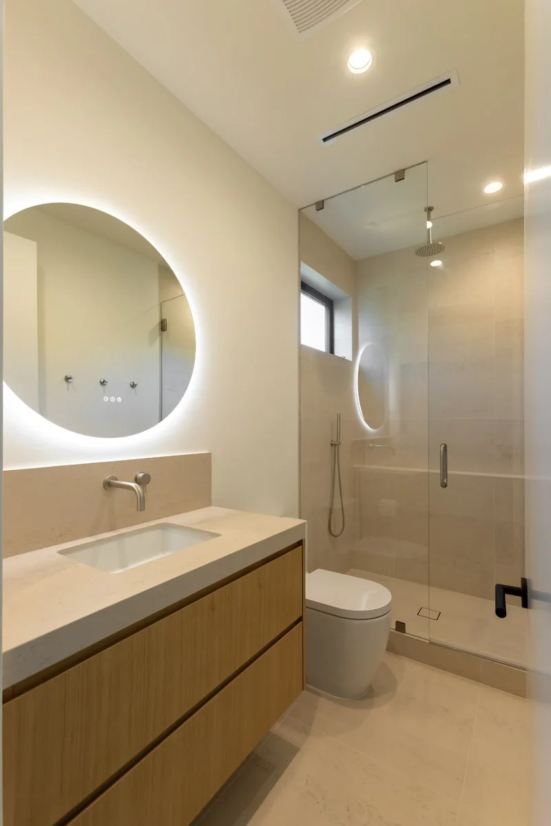 Modern Mar Vista bathroom remodel with floating light wood vanity, integrated stone countertop, round backlit mirror, walk-in glass shower, and minimalist contemporary fixtures in a custom home.