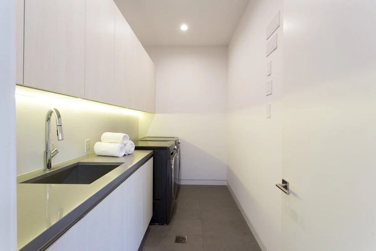Minimalist laundry room with flat-panel cabinetry, under-cabinet LED lighting, integrated utility sink, quartz countertop, and stacked front-load washer and dryer.