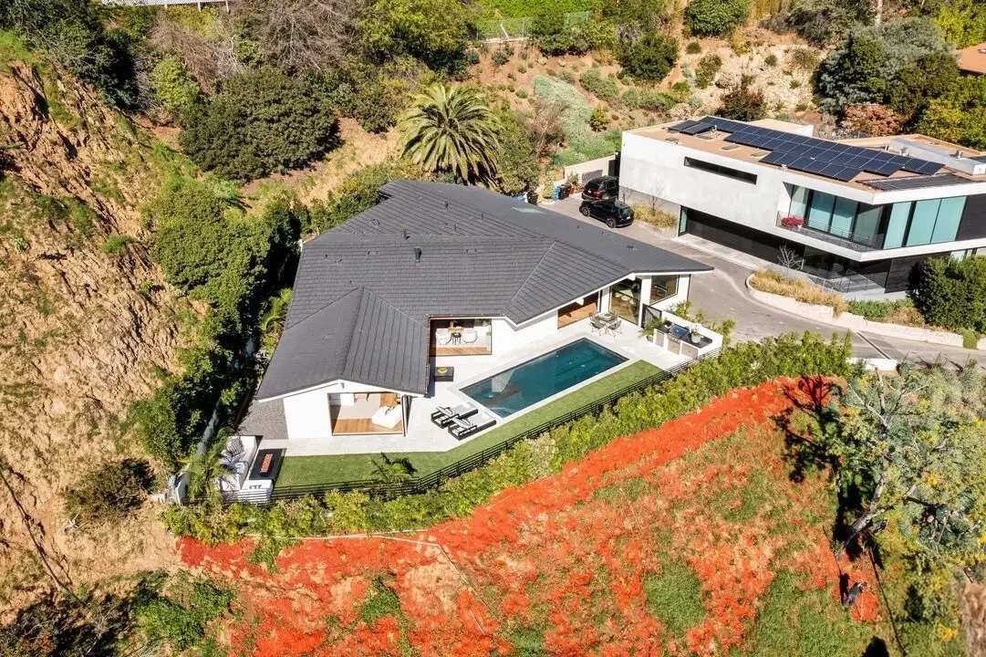 A modern house with a dark grey roof, swimming pool, and outdoor lounging area, located on a hillside with surrounding trees and vegetation.