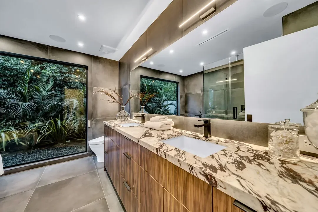 Modern bathroom with marble countertop, wooden cabinet, large mirror, glass-enclosed shower, and windows overlooking indoor plants.