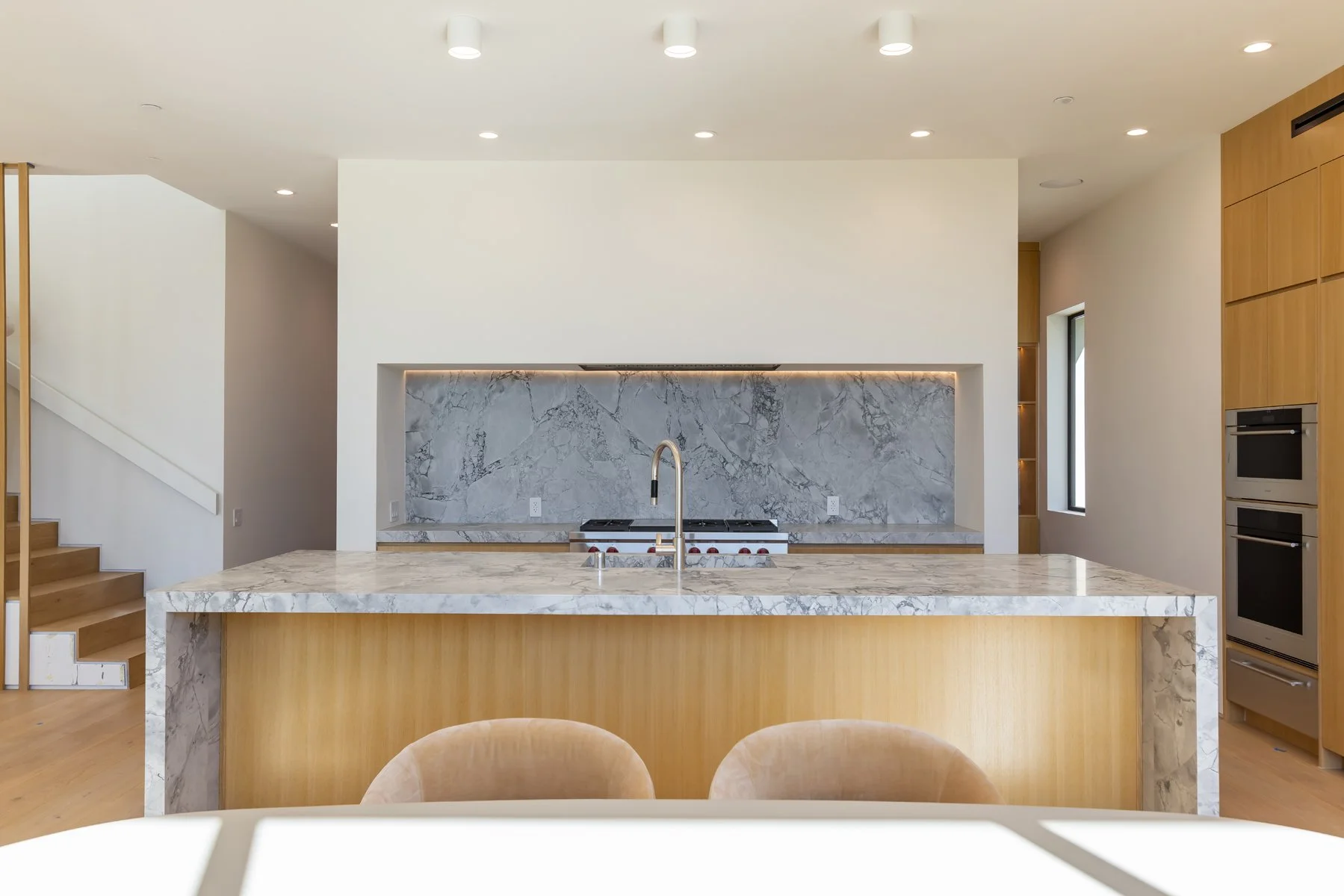 Minimalist Mar Vista kitchen with a marble waterfall island, integrated gas cooktop, warm wood cabinetry, and recessed lighting in a custom modern home.