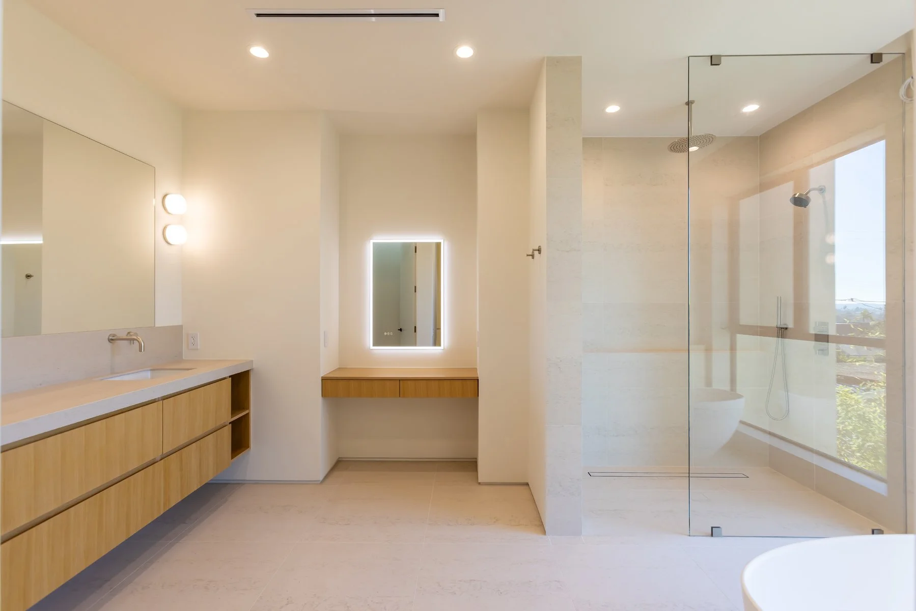 Modern Mar Vista primary bathroom with floating light wood vanity, integrated makeup station with backlit mirror, large glass walk-in shower, and minimalist contemporary finishes by Heart Construction.