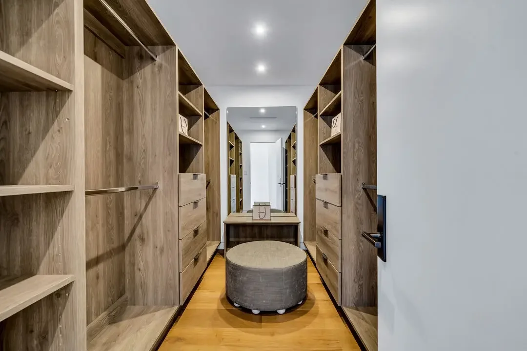 Walk-in closet with wooden shelves and drawers, a round ottoman in the center, and a mirror at the end, illuminated by recessed ceiling lights.