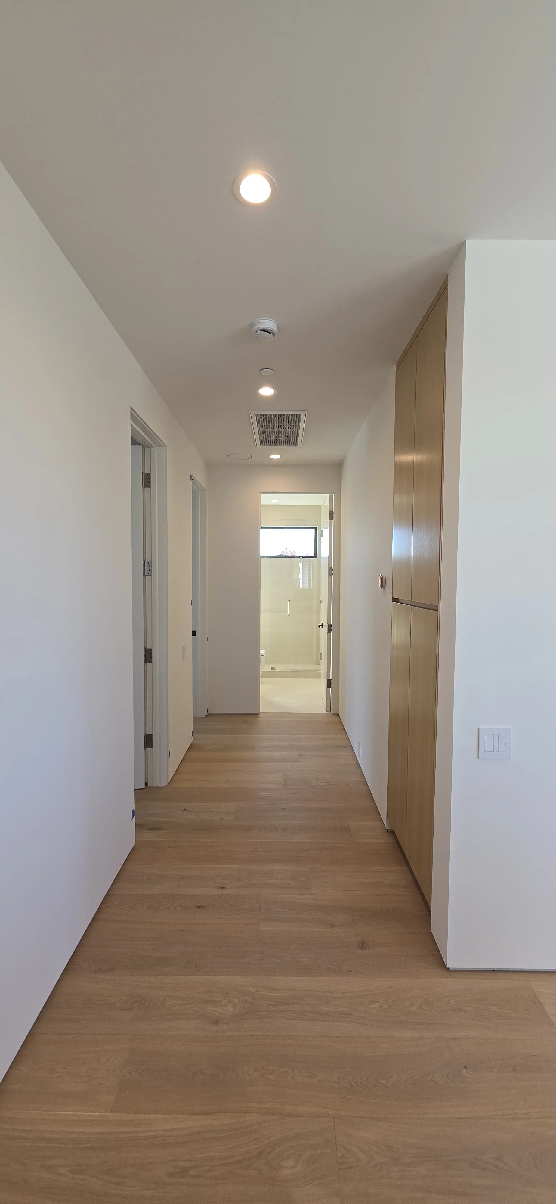 Clean modern hallway with wide-plank oak flooring, recessed ceiling lighting, and flush wood cabinetry, leading to a light-filled bathroom in a Mar Vista custom home by Heart Construction.