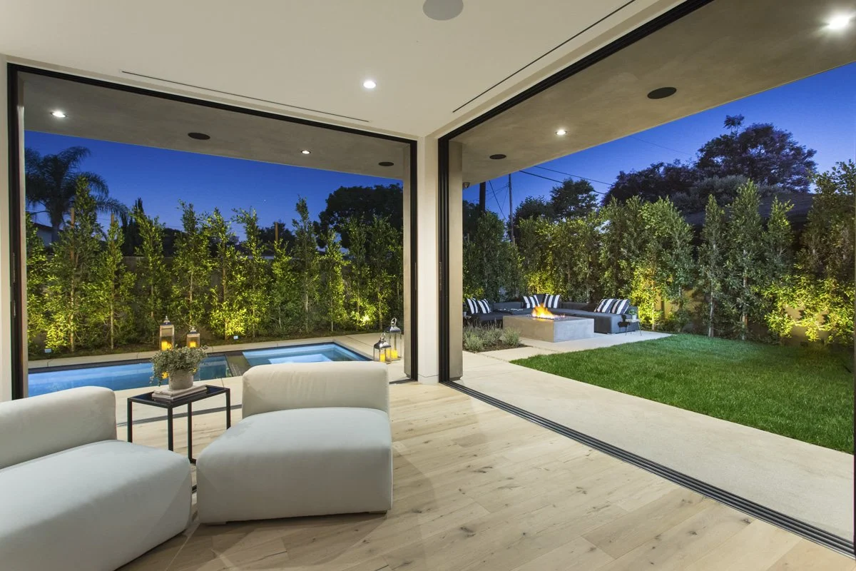 Modern great room with corner sliding glass walls opening to lawn and pool, low-profile lounge seating, recessed lighting, and continuous oak flooring.