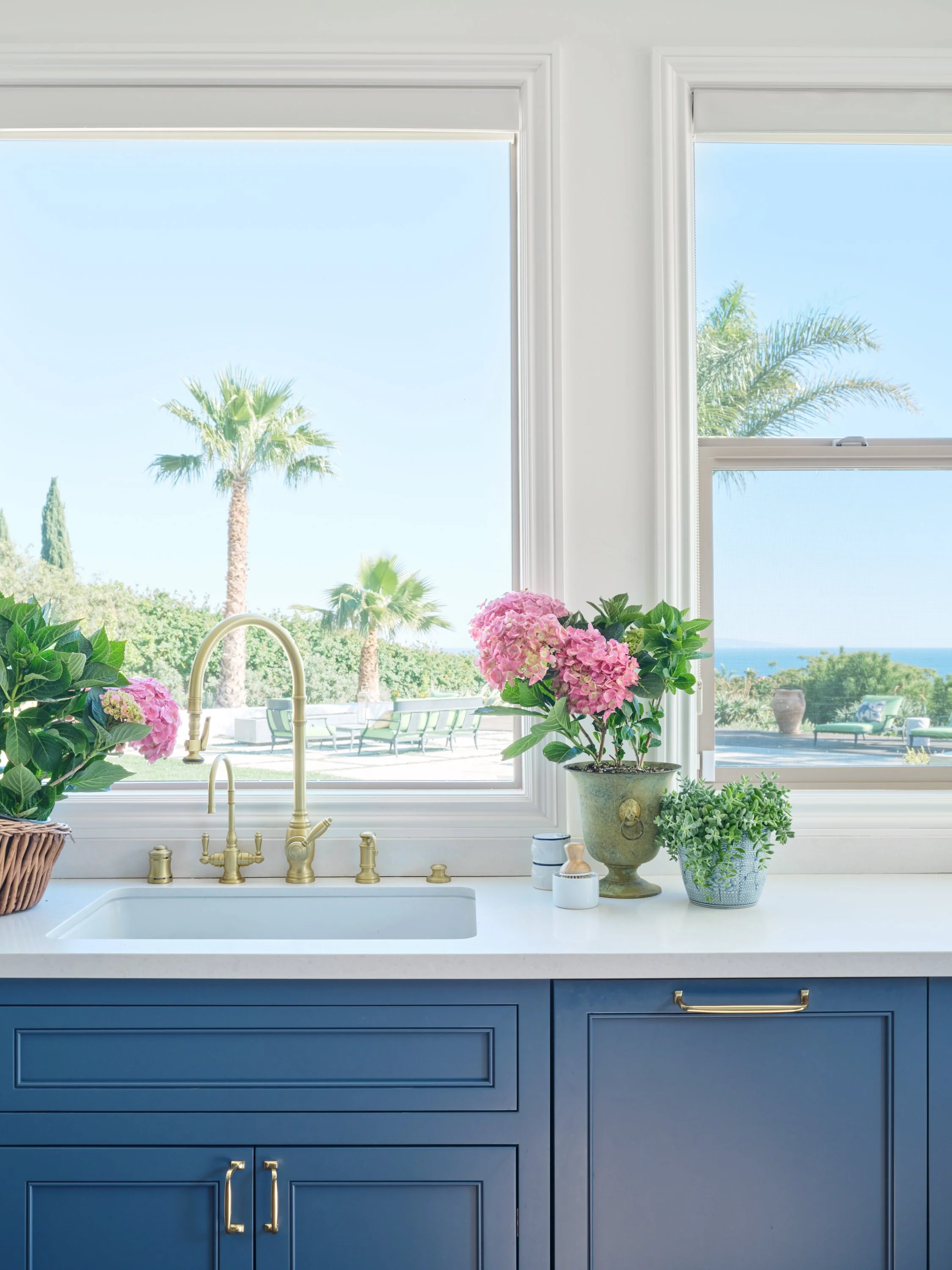 Malibu kitchen with ocean view, white countertops, blue cabinetry, brass faucet, and palm trees visible through large windows