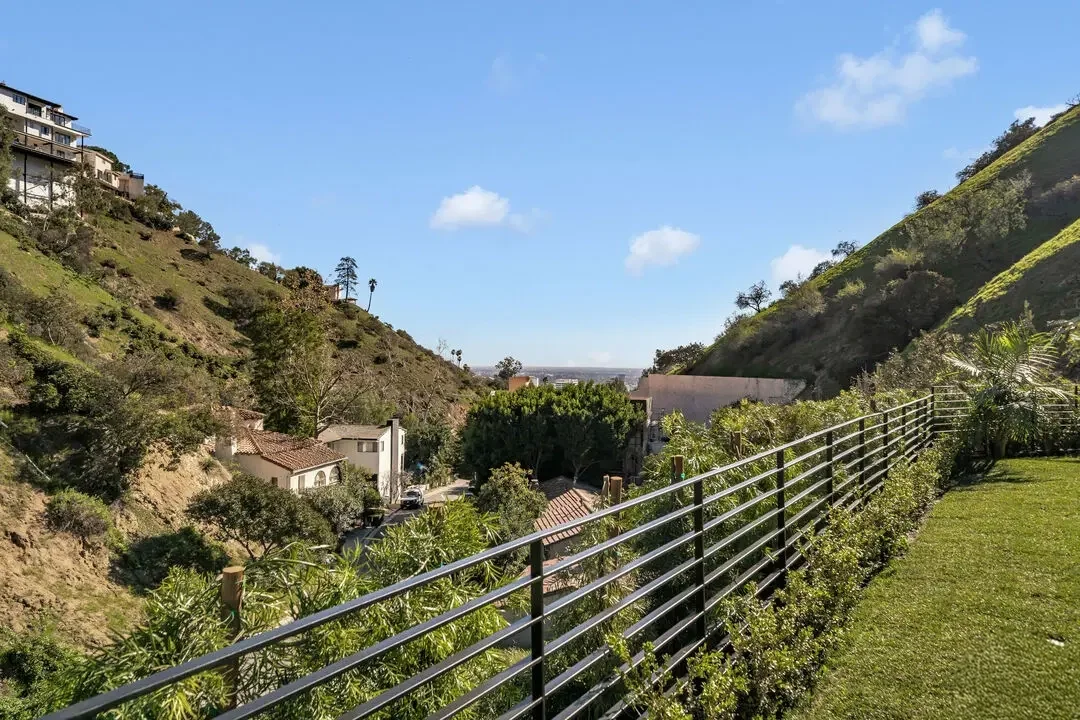 View of a hillside neighborhood with houses, trees, and green slopes, seen from a fenced balcony or patio area with manicured grass and hedges, under a blue sky with a few clouds.