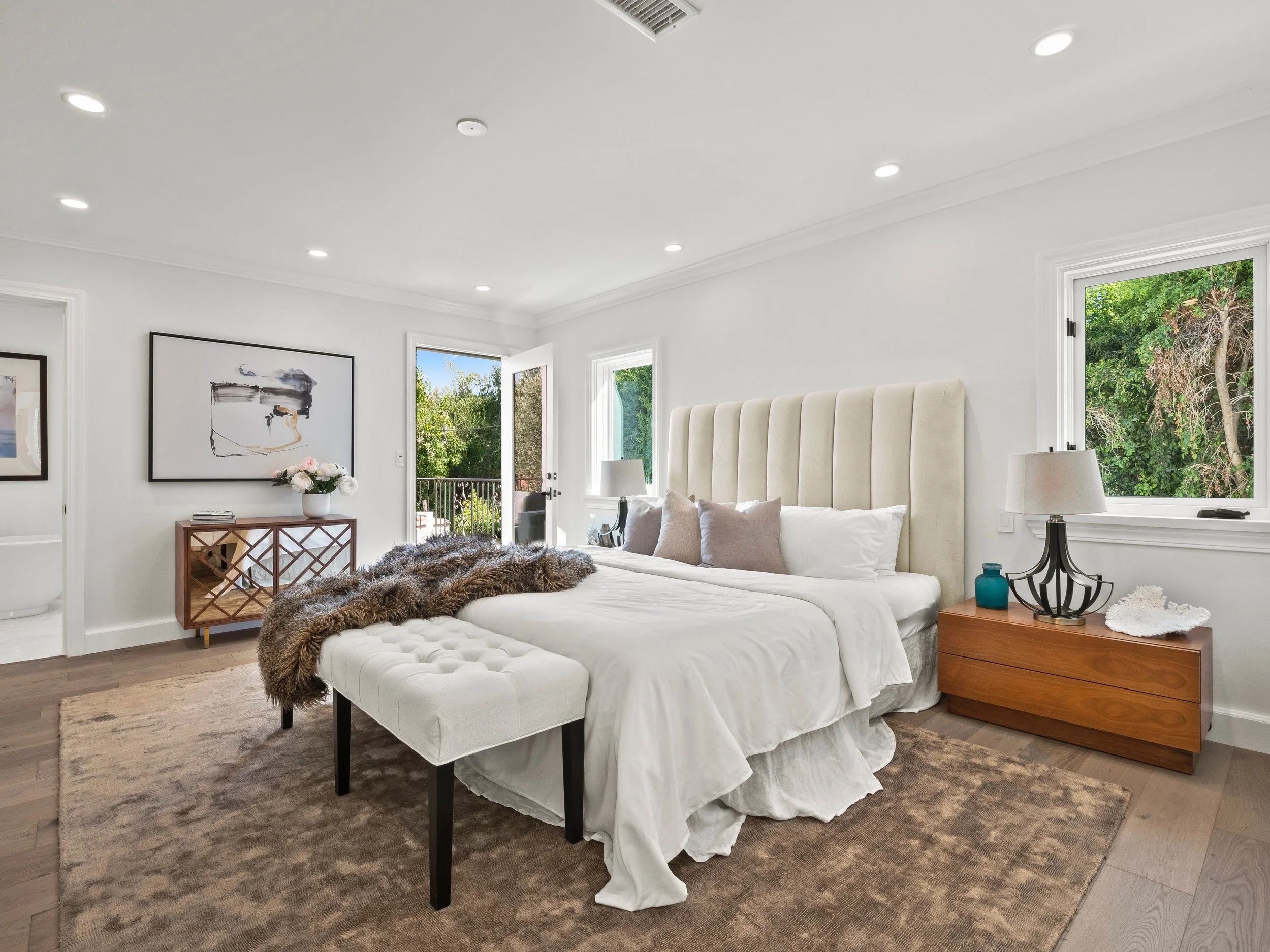 Hancock Park primary bedroom with tall upholstered headboard, neutral layered bedding, wood nightstands, and French doors opening to a private balcony.
