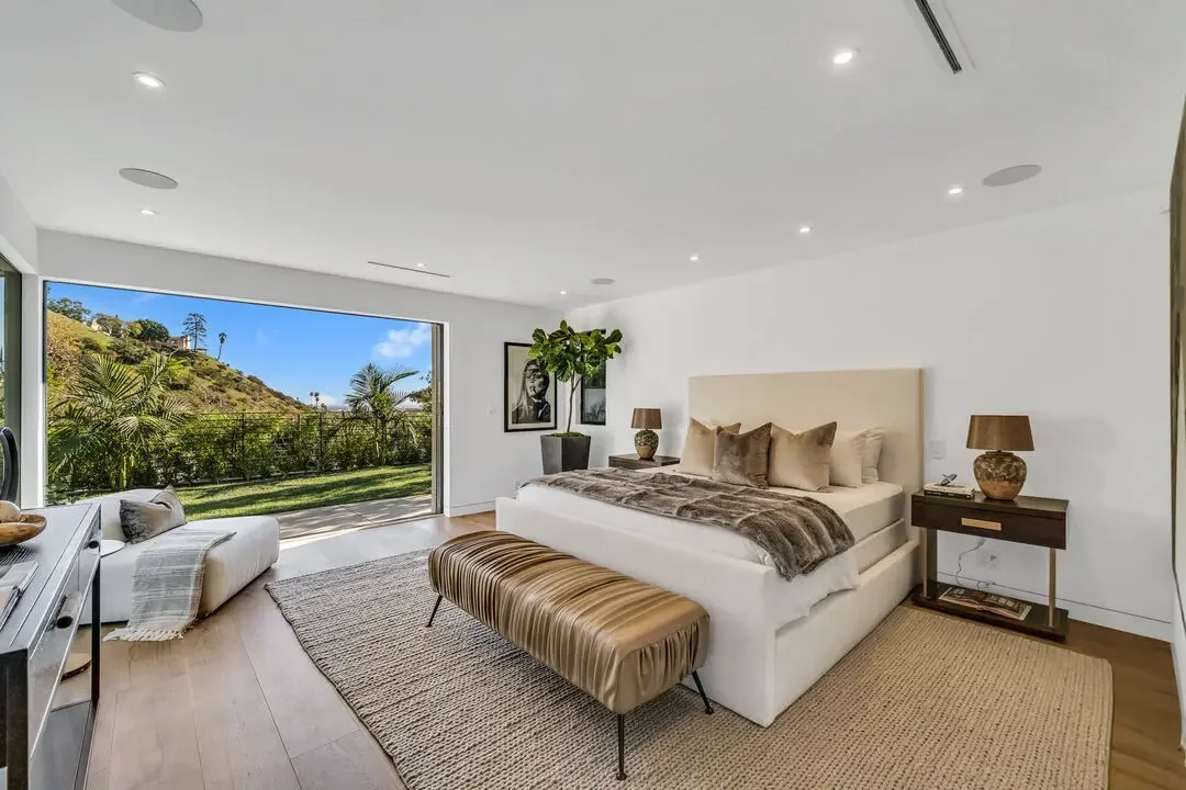 Modern bedroom with large window view of green landscape, outdoor trees, and hills, featuring a white bed with beige pillows, brown throw blanket, wooden bedside tables with lamps, a striped bench at the foot of the bed, and art on the wall.