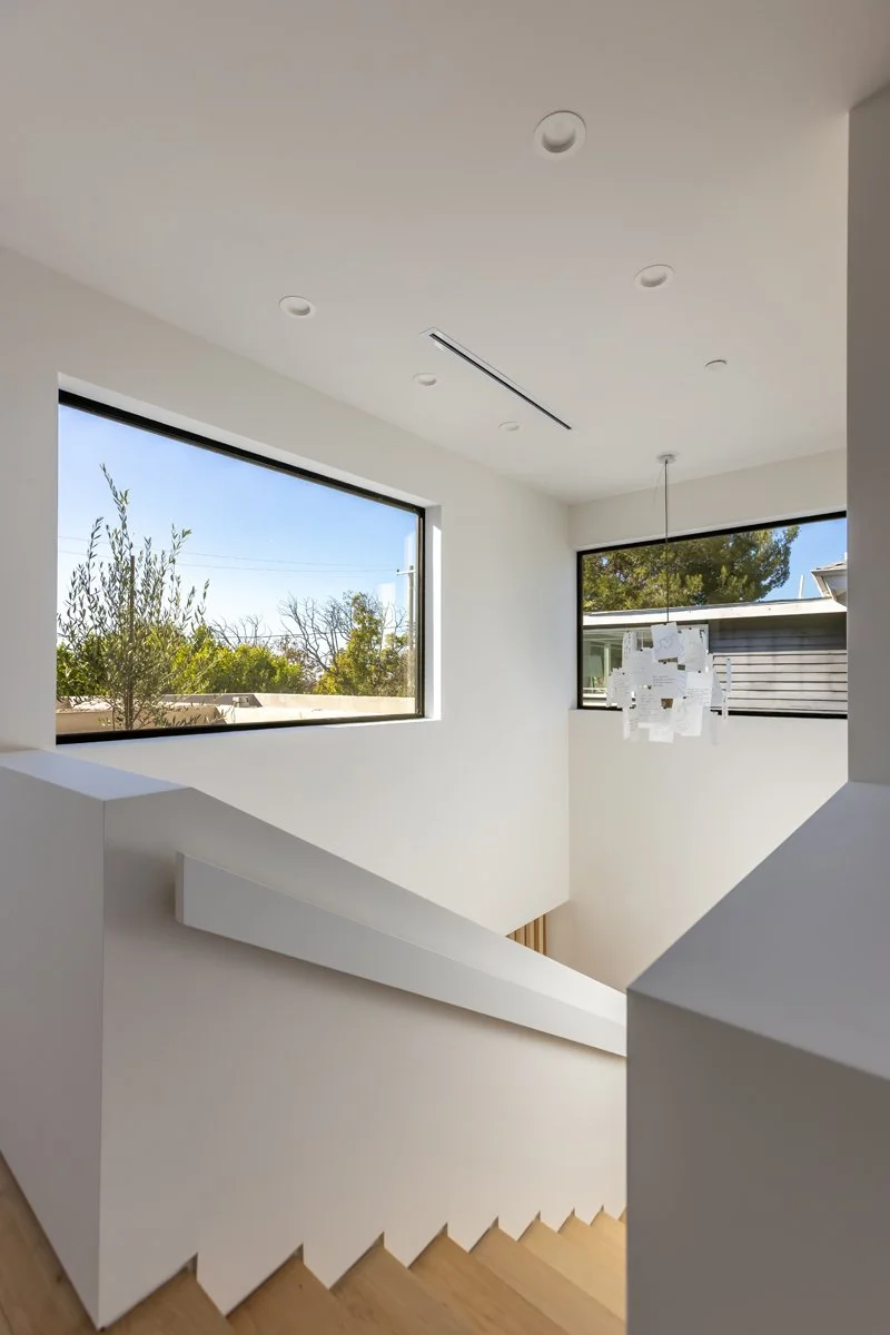 Minimalist stairwell with light oak treads, white walls, recessed lighting, and large picture windows bringing natural light into a modern Mar Vista custom home by Heart Construction.