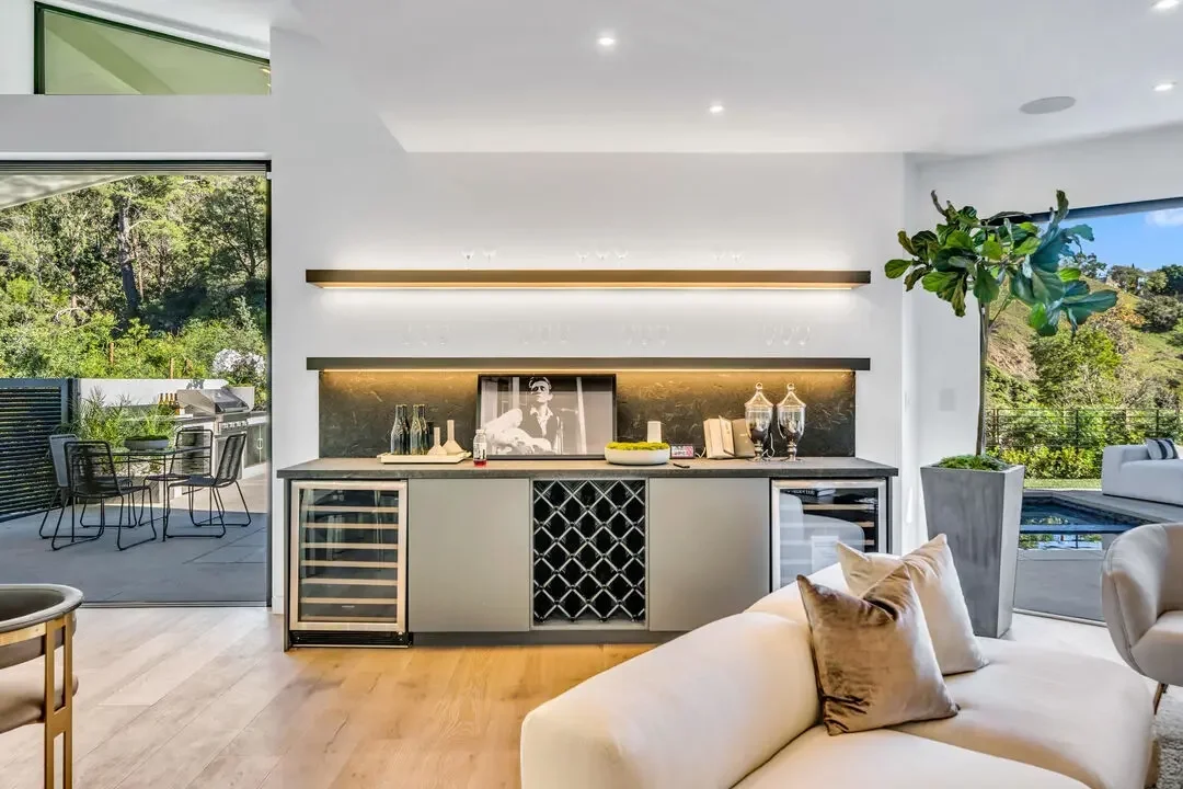 Modern living room with a built-in bar, large window with outdoor patio and greenery, and a potted plant.