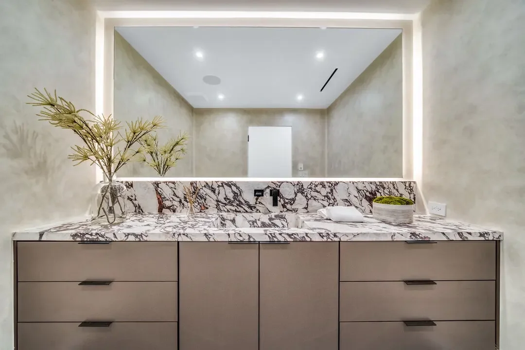 Modern bathroom vanity with a marble countertop, large mirror, and minimal decor, including a vase with green foliage and a small bowl with moss.