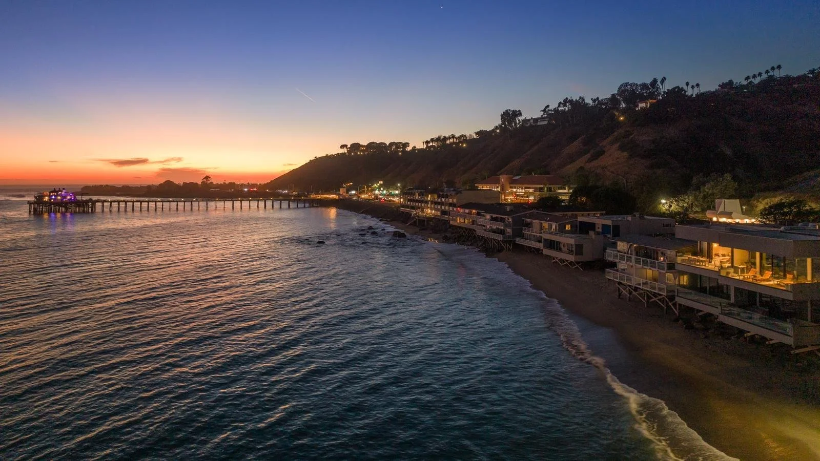 Malibu coastline at sunset with oceanfront homes along the sand, hillside backdrop, and illuminated pier extending into the Pacific.