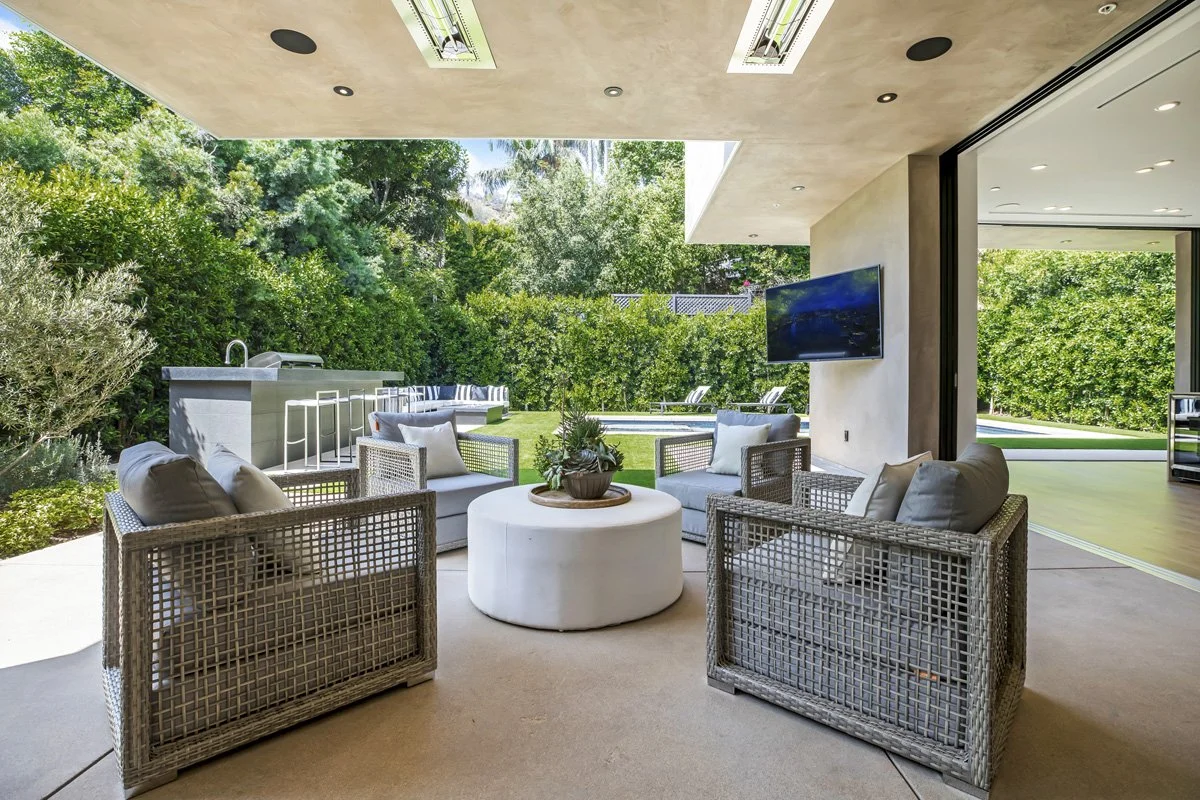 Covered outdoor living area in Beverly Hills with lounge seating, retractable glass wall, and view of custom pool and lawn.