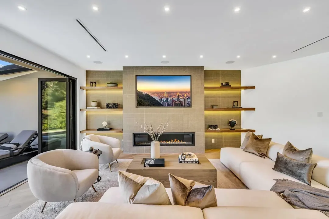 Modern living room with a large white sofa, beige armchair, and a gray lounge chair, with a wall-mounted TV, fireplace, and wooden shelves, opening to a balcony with outdoor seating, and decorated with cushions, vases, and books.