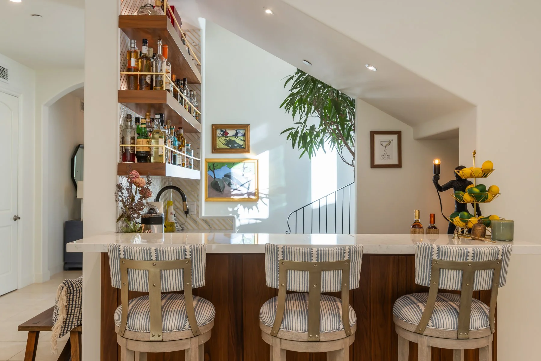 Built-in home bar in a custom Brentwood residence featuring a marble countertop, walnut cabinetry, open wood shelving with liquor display, upholstered bar stools, and natural light from a nearby stairwell.