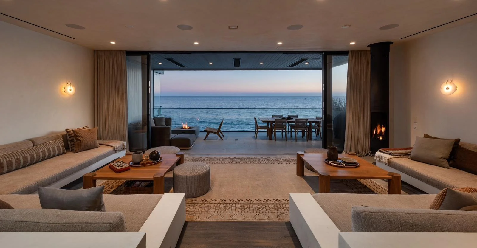 Malibu oceanfront living room with symmetrical seating, fireplace accents, and sliding glass doors opening to a terrace overlooking the Pacific at sunset.