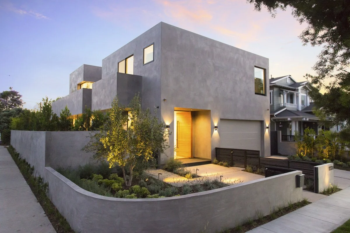 Venice modern custom home corner elevation at dusk with stacked plaster volumes, warm wood entry door, uplighted olive trees, and concrete perimeter wall