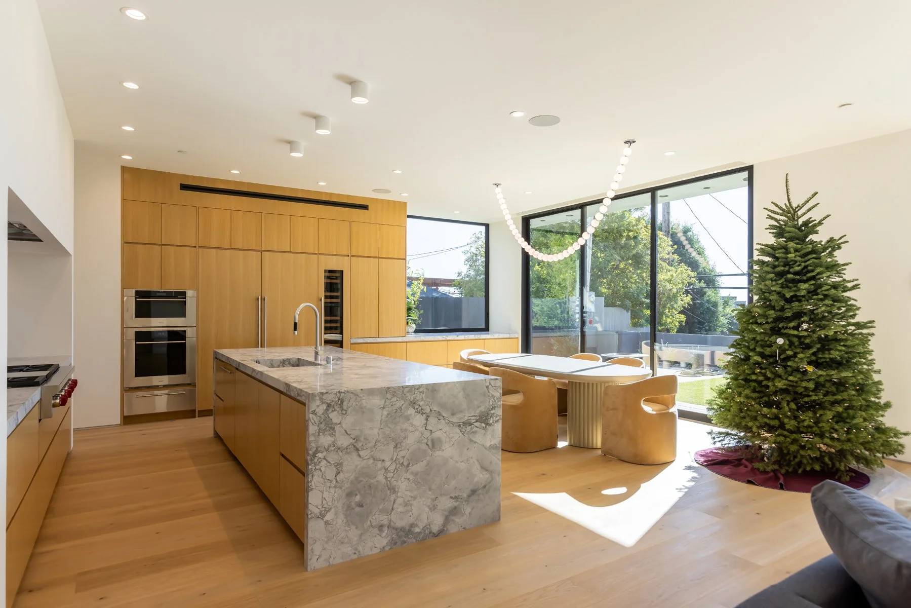 Modern kitchen and dining area in a custom Mar Vista home featuring a marble waterfall island, warm wood cabinetry, and floor-to-ceiling glass doors with natural light.