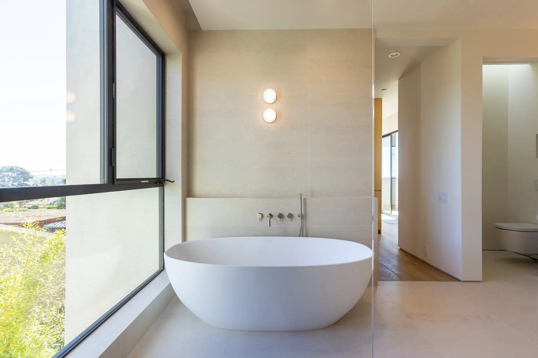 Minimalist freestanding soaking tub set against large-format stone tile, floor-mounted tub filler, and expansive window in a modern Mar Vista primary bathroom remodel by Heart Construction.