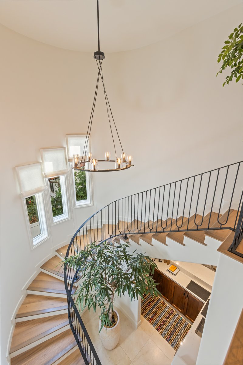 Curved staircase in a custom Brentwood home featuring wood treads, a black iron railing, sculptural chandelier, tall windows, and a central indoor tree bringing in natural light.