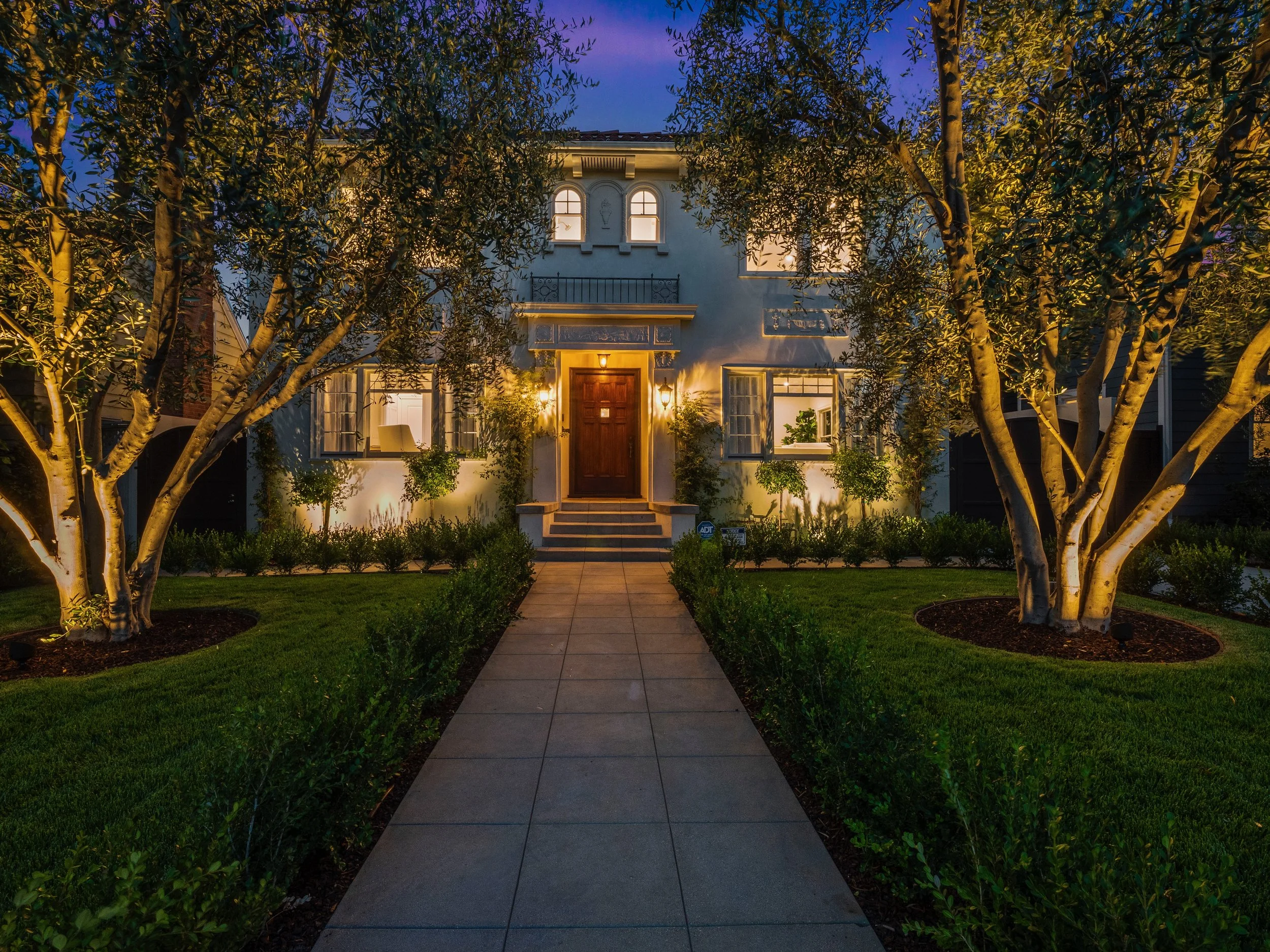 Calabasas custom home exterior at dusk, featuring symmetrical Mediterranean architecture, warm landscape lighting, arched windows, and a manicured front walkway framed by mature trees.