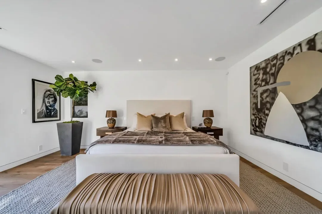 A modern bedroom with a large bed, beige pillows, and a fur blanket, flanked by two wooden nightstands with brown lamps, a large abstract painting on the right, a black-and-white portrait on the left, and a potted plant with broad green leaves in a tall gray planter.