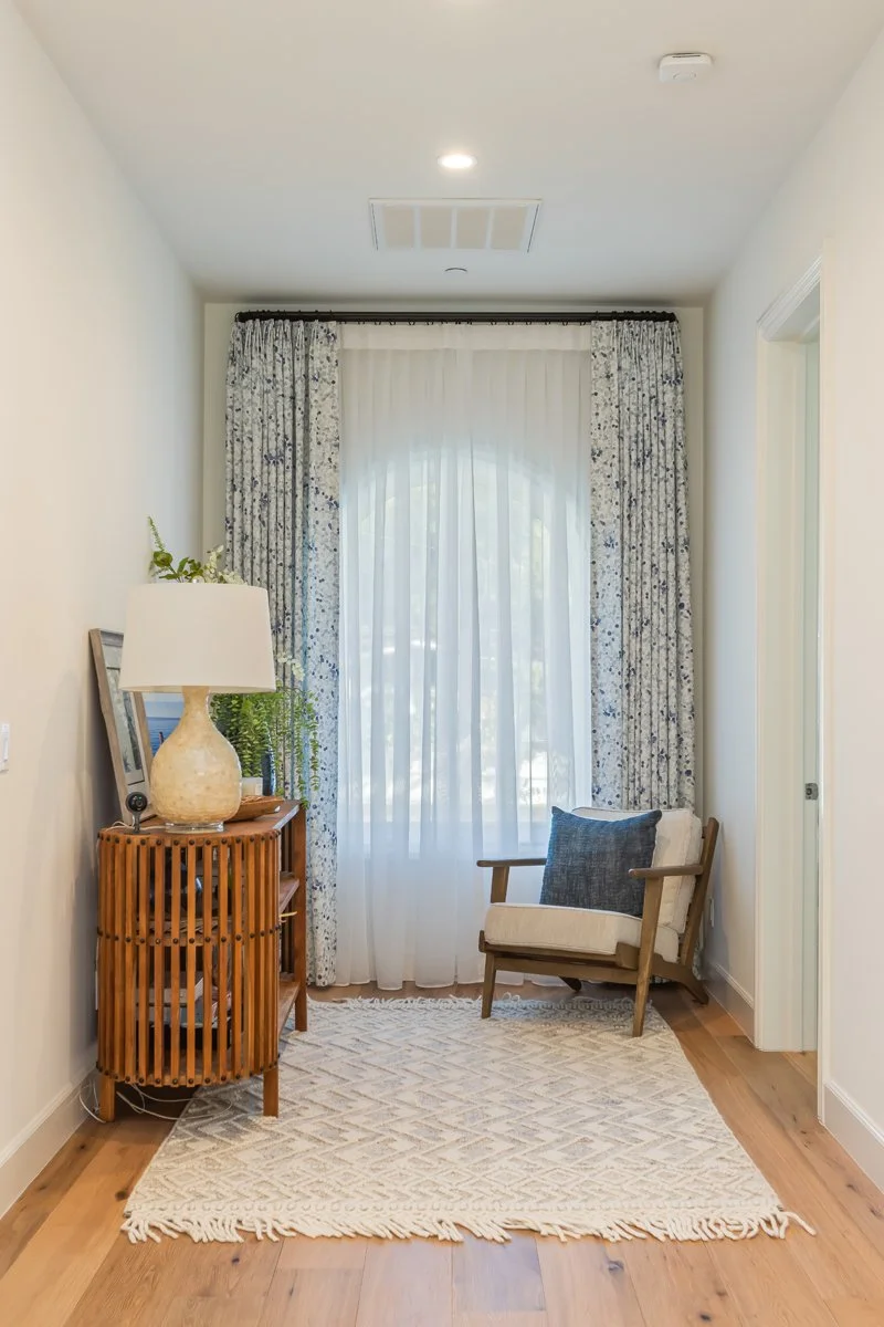 Cozy sitting nook in a custom Brentwood home featuring a wood lounge chair, patterned area rug, natural wood flooring, soft window drapery, and warm ambient lighting.