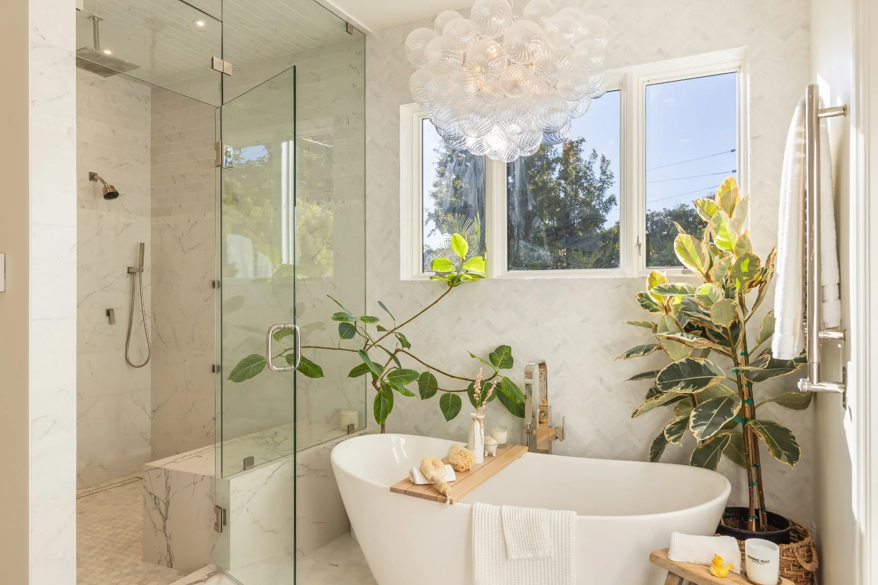 Freestanding soaking tub in a bright Brentwood primary bathroom with glass-enclosed marble shower, herringbone tile walls, and natural light from large windows.