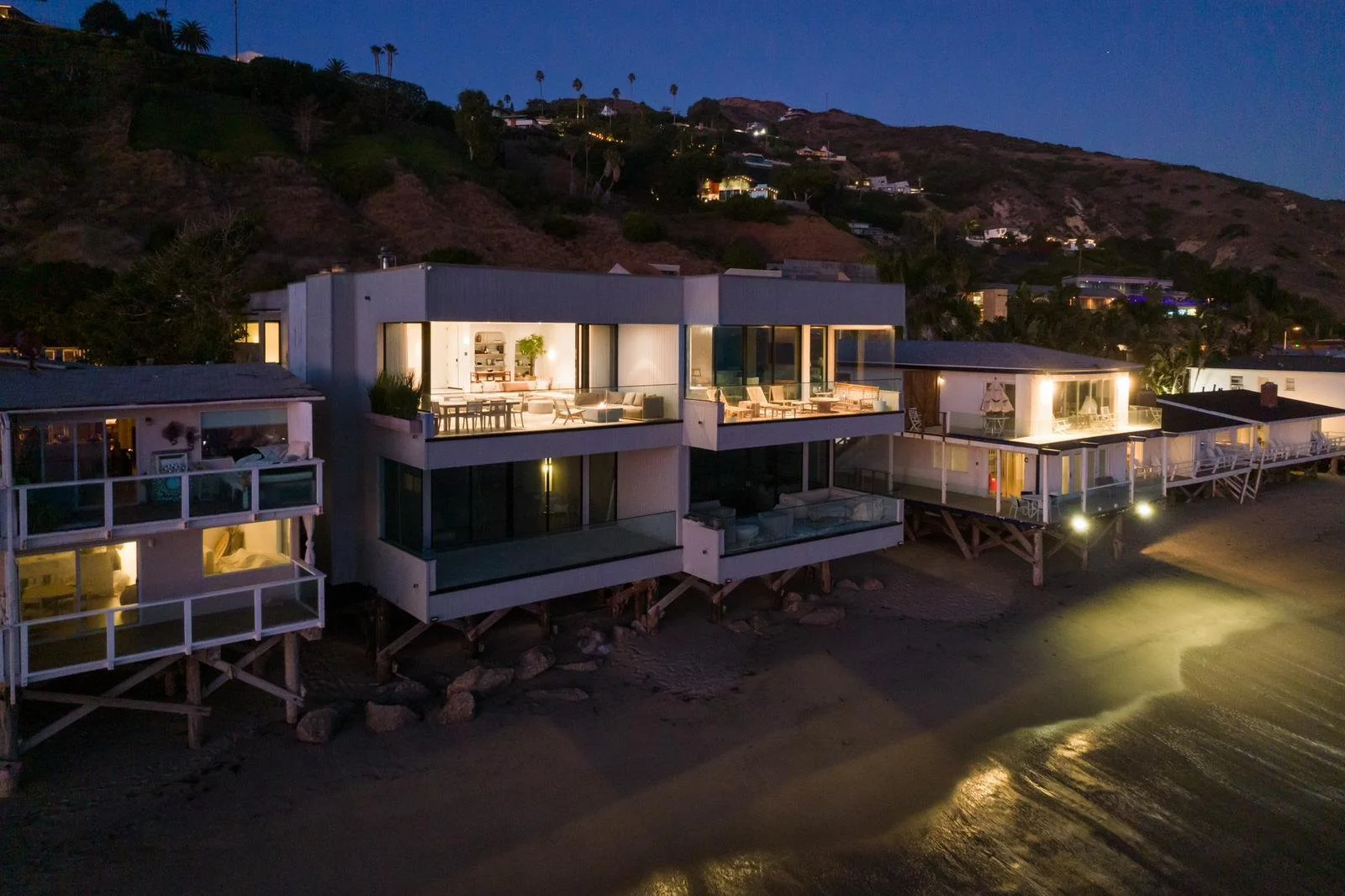 Malibu contemporary oceanfront home at dusk, elevated over the sand with expansive glass walls, illuminated terraces, and hillside backdrop.