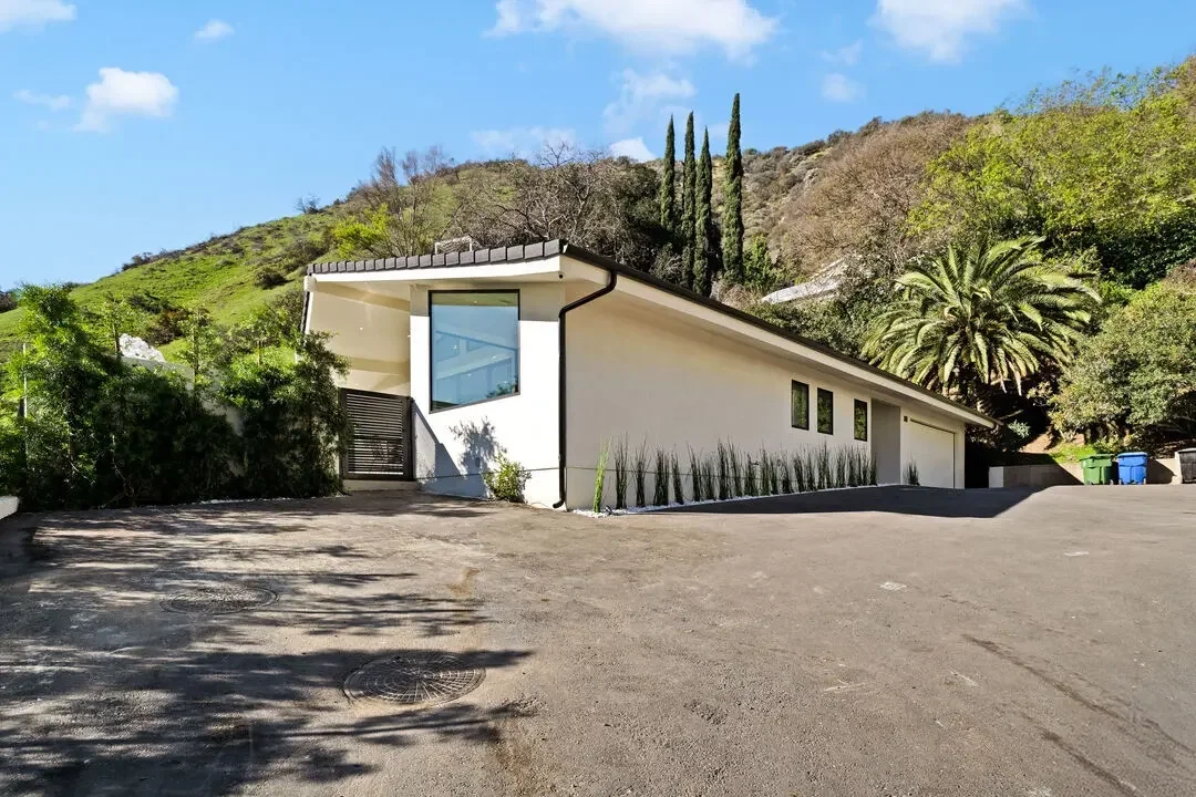 A modern, single-story house with white exterior walls and black trim, situated on a slightly inclined driveway with parking space. The house is surrounded by lush greenery, including trees and bushes, with a hillside in the background and a partly cloudy blue sky overhead.