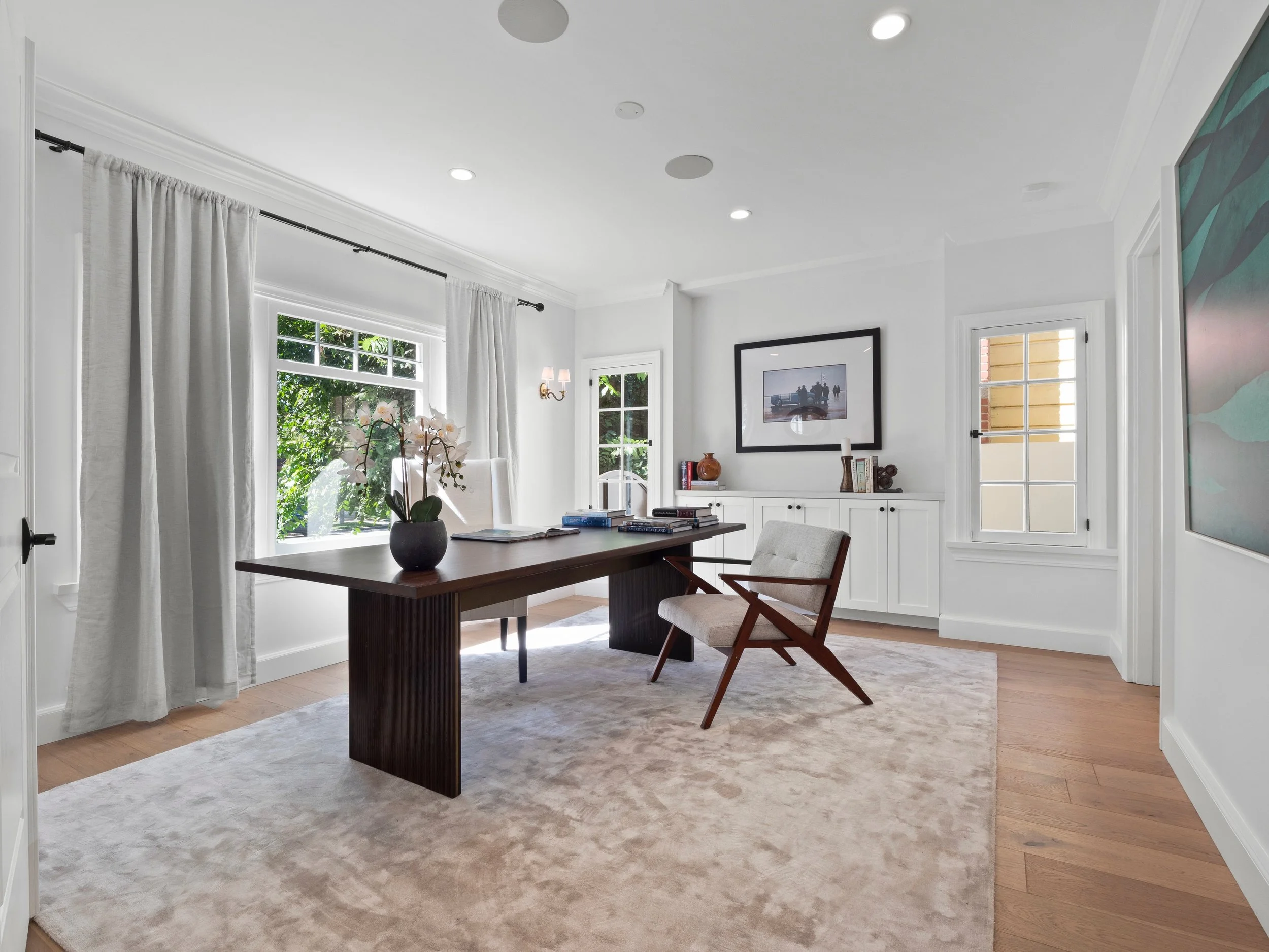 Hancock Park home office with custom millwork, dark wood desk, upholstered accent chair, soft neutral rug, and large windows bringing in natural light.