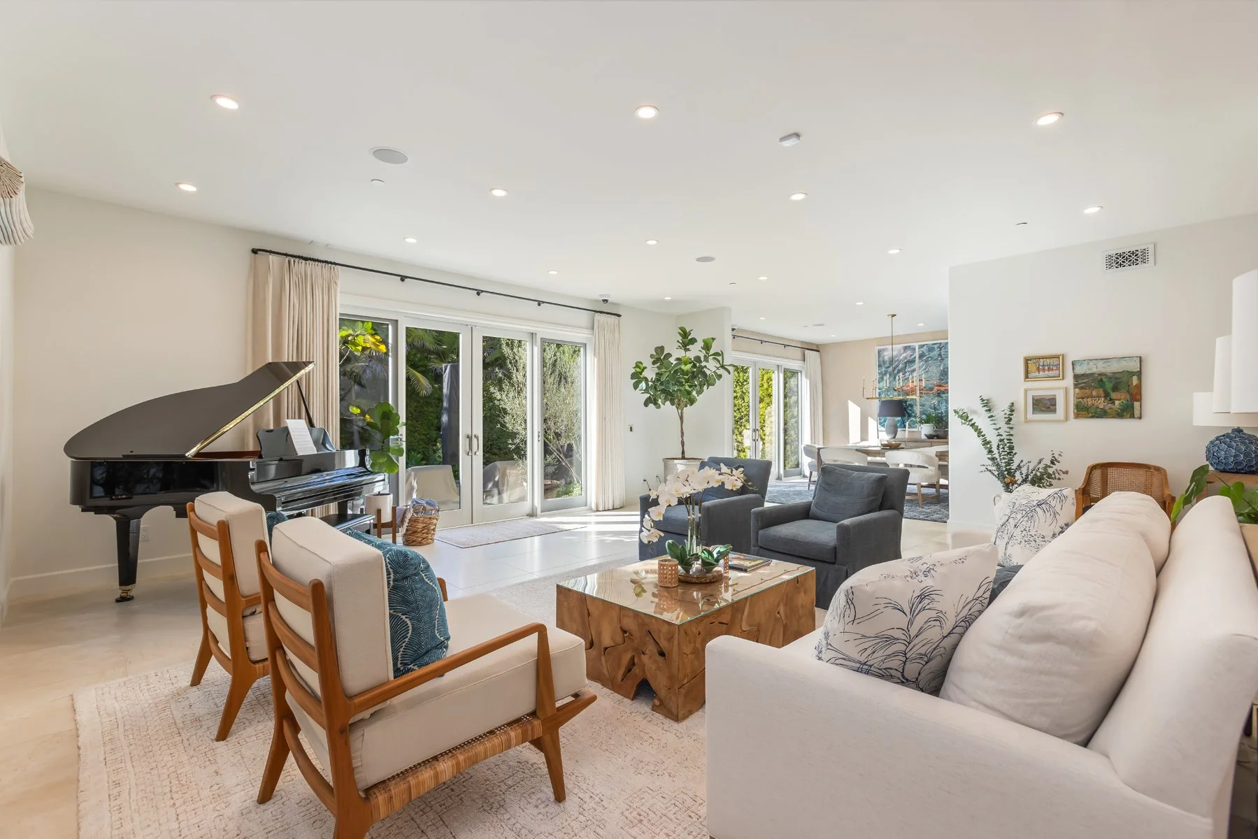 Open-concept living room in a custom Brentwood home featuring a grand piano, layered seating, light stone floors, recessed lighting, and large glass doors opening to the backyard.