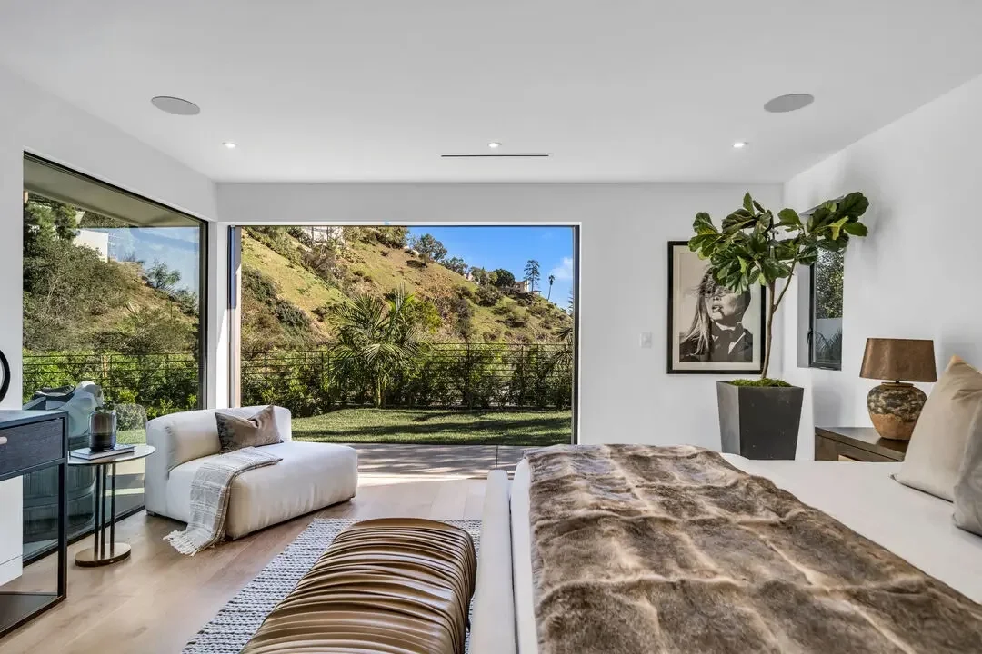 Modern bedroom with large glass sliding doors displaying a green hillside landscape, featuring a bed with fur blanket, a plush armchair, a large potted plant, framed black-and-white portrait, and contemporary decor.