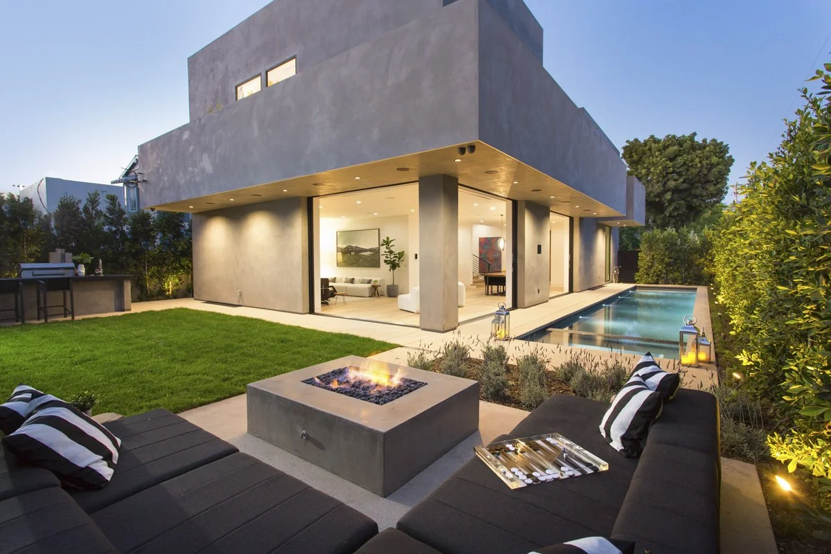 Venice modern custom home exterior at twilight with stacked plaster volumes, wraparound glass walls, illuminated soffits, and lap pool anchoring the yard.