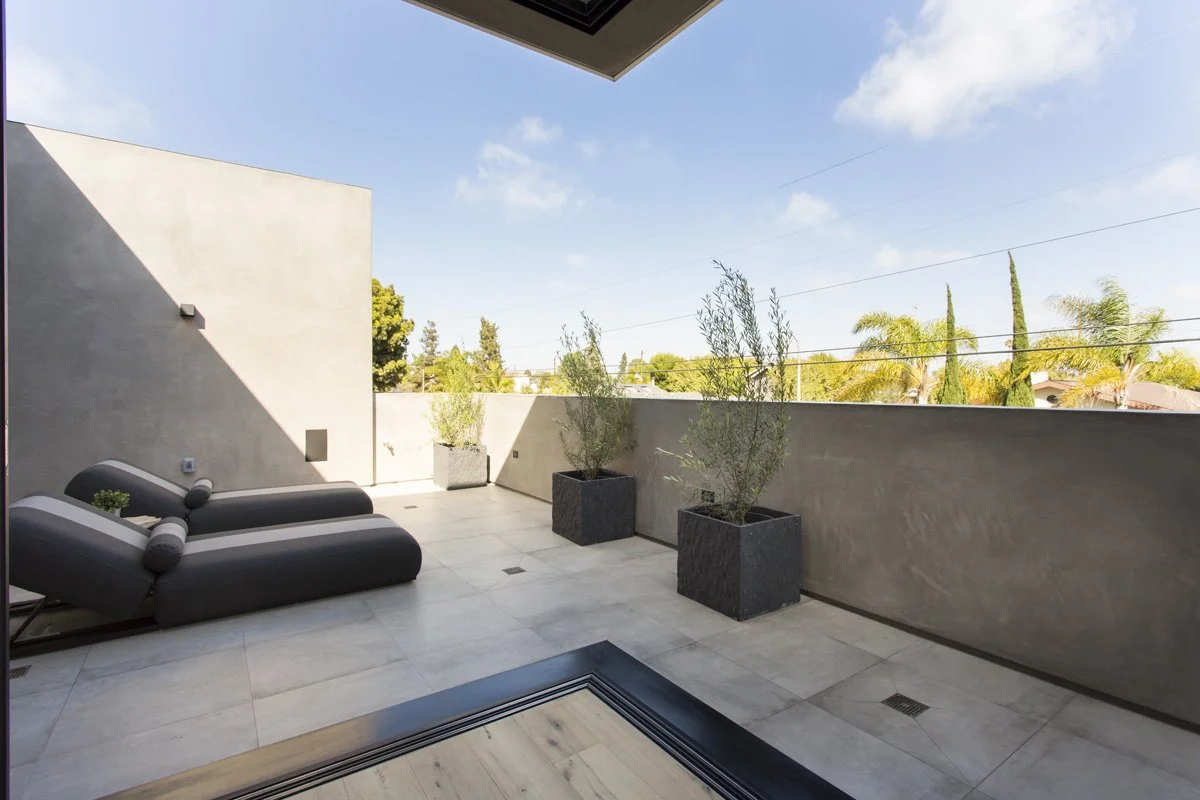 Private rooftop terrace in Venice custom home with oversized chaise loungers, large-format porcelain pavers, built-in drainage, and skyline views above tree canopy.