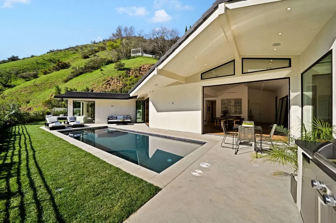 Modern suburban house with a rectangular swimming pool, outdoor seating area, and hillside with greenery in the background.