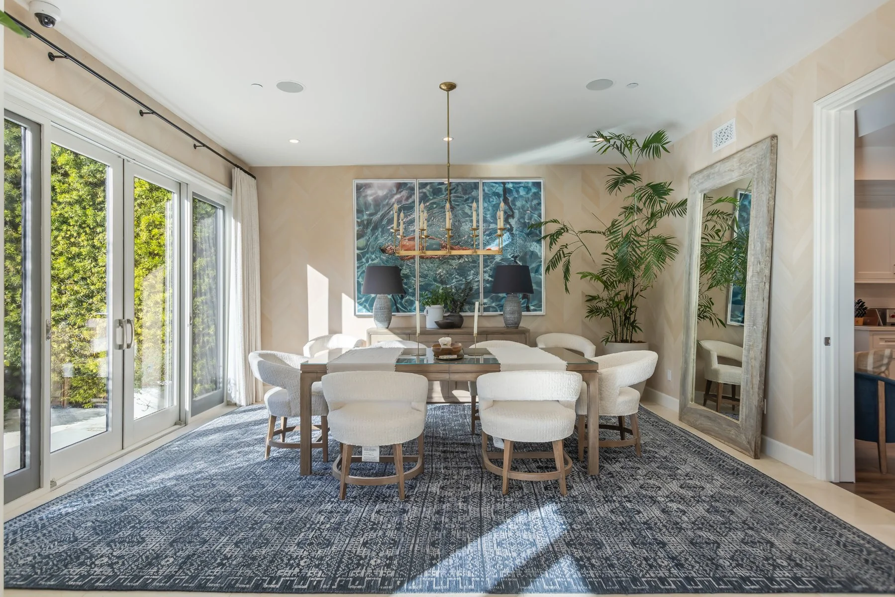 Dining room in a custom Brentwood home featuring a wood dining table, upholstered chairs, statement chandelier, large artwork, and glass doors opening to the garden.