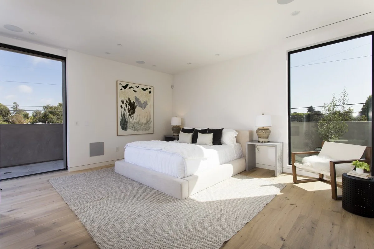 Primary bedroom in Venice modern residence with low upholstered platform bed, expansive corner glazing, natural oak floors, and private balcony access.