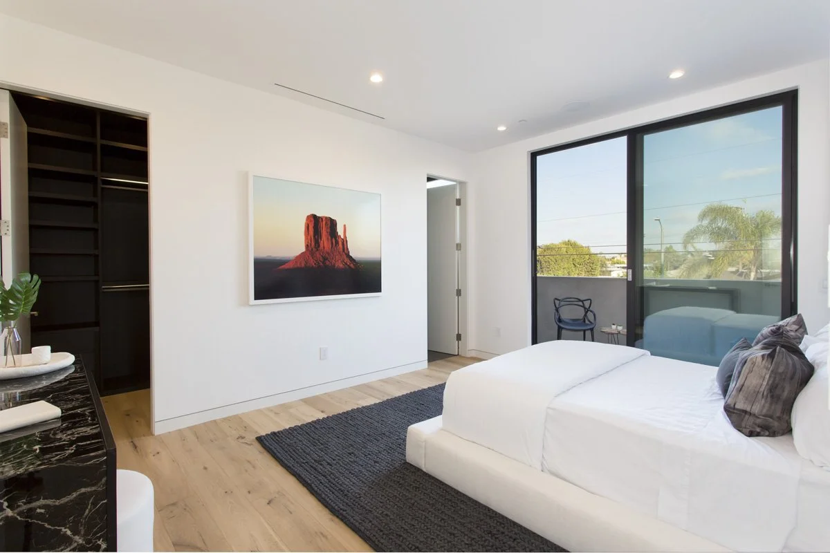 Contemporary bedroom with upholstered platform bed, wide-plank oak flooring, large sliding glass door opening to private balcony, and minimalist white walls.